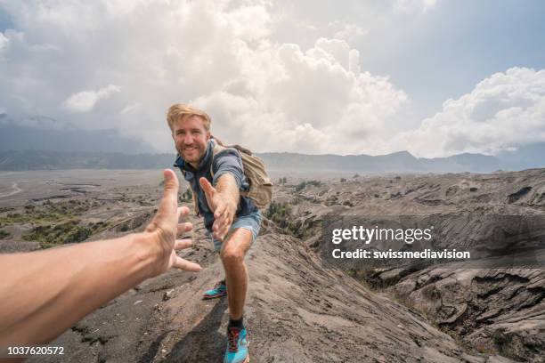 young man hiking, pulls out hand to reach the one of teammate. a helping hand to reach the top of crater volcano. bromo volcano region in indonesia, asia - leading the way forward stock pictures, royalty-free photos & images