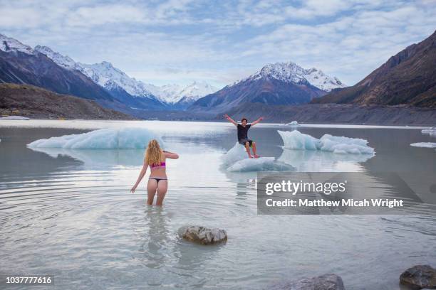 friends walk into the icy waters of the glacial lakes of mount cook national park. a man sits on a floating iceberg. - new zealand travel stock pictures, royalty-free photos & images