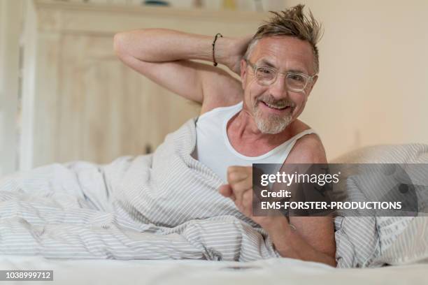 happy senior adult man in his early 60s with grey beard and eyeglasses chilling in bed during day. - ascella foto e immagini stock