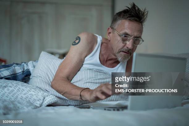 senior active man in his 60s with greying hair and eyeglasses in tank top sleepless in bed while night using his laptop. - addiction mobile and laptop stock-fotos und bilder