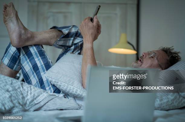 unhealthy senior man in his 60s with greying beard sleepless in bed while night using laptop and smart phone. - 不穏状態 ストックフォトと画像