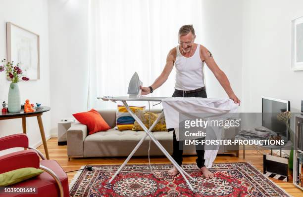 happy, multi-tasking senior ironing his shirt in living room at home. - bügeln stock-fotos und bilder