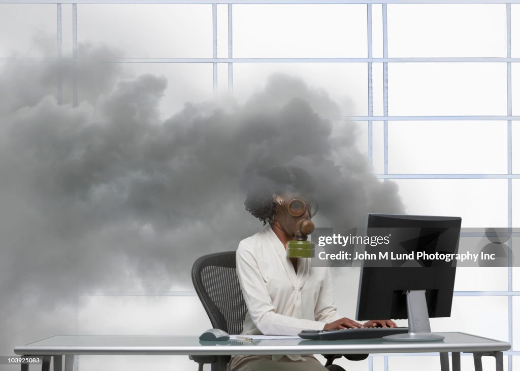 Black businesswoman wearing gas mask at smoking computer