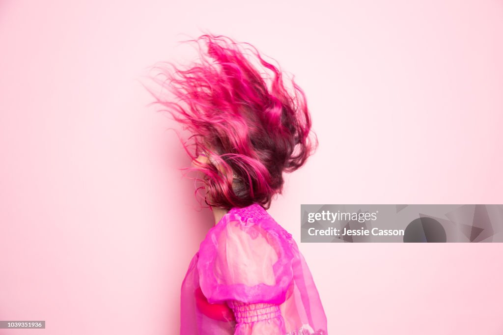 A woman with pink hair throws her head back so that her hair covers her face in front of a pink background