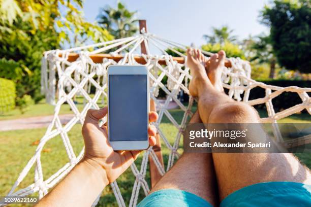 personal perspective view of a man swinging in hammock and using smart phone - smartphone strand stock-fotos und bilder