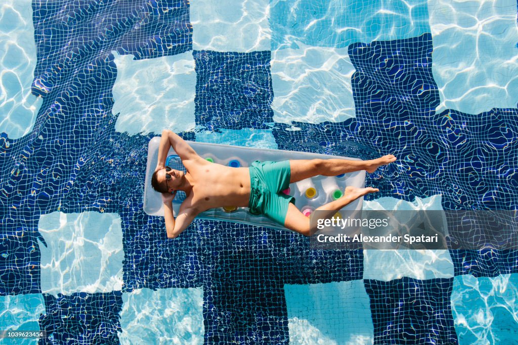 High angle view of a young man in sunglasses sunbathing in swimming pool on inflatable pool raft