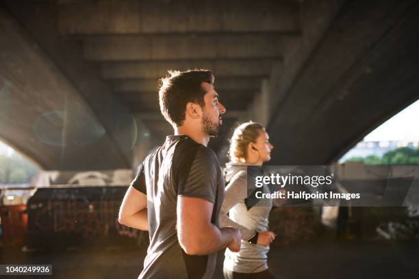 young sporty man and woman with earphones running under the bridge outside in a city. - joggen stockfoto's en -beelden