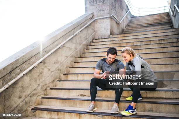 young sporty man and woman with earphones sitting on stairs outside in a city, using smart watch. - england slovakia stock pictures, royalty-free photos & images
