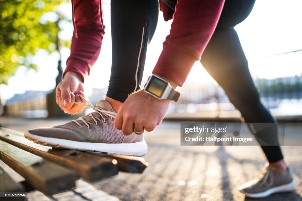 Young sporty man with smart watch tying shoelaces on a bench outside in a city at sunset.