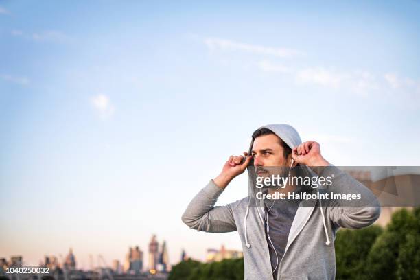 young sporty man with earphones putting a hood on his head outside in a city, listening to music. - england slovakia stock pictures, royalty-free photos & images