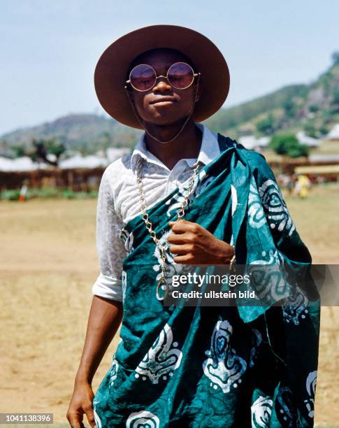 Junger Afrikaner eigenwillig modebewusst mit rundem Hut, einer Riesenbrille, langer Schmuckhalskette und weissem Hemd, in ein traditionell buntes...