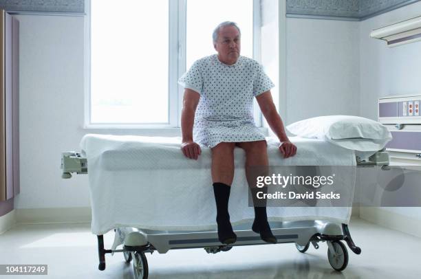 senior man sitting on bed in hospital room - hospital gown stock photos et images de collection