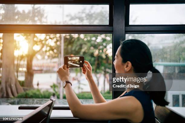 young woman photographing city scene by the window with smartphone during sunset while riding on public transportation in city - straßenbahnstrecke stock-fotos und bilder