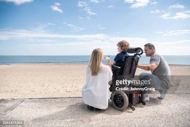 ouders crouching door zoon in rolstoel op strand - wheelchair stockfoto's en -beelden