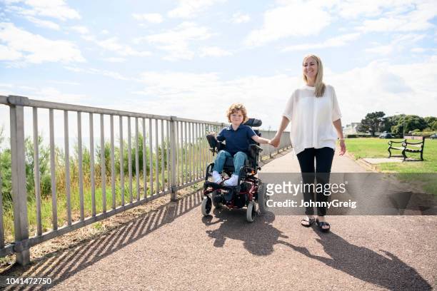 disabled boy in wheelchair holding mother's hand on path - special needs children stock pictures, royalty-free photos & images