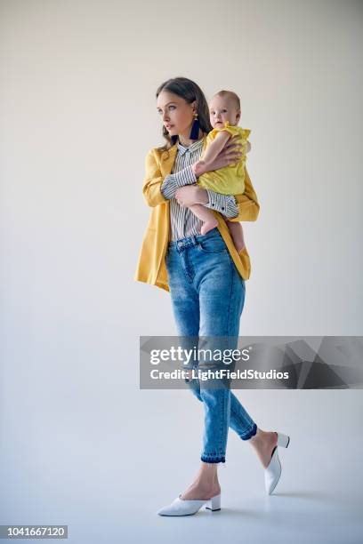 full length view of stylish young mother carrying adorable baby daughter and walking on grey