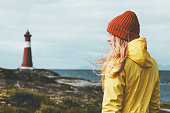 Woman sightseeing lighthouse sea landscape in Norway Travel Lifestyle concept scandinavian vacations outdoor. Blonde girl hair on wind wearing orange hat and yellow raincoat