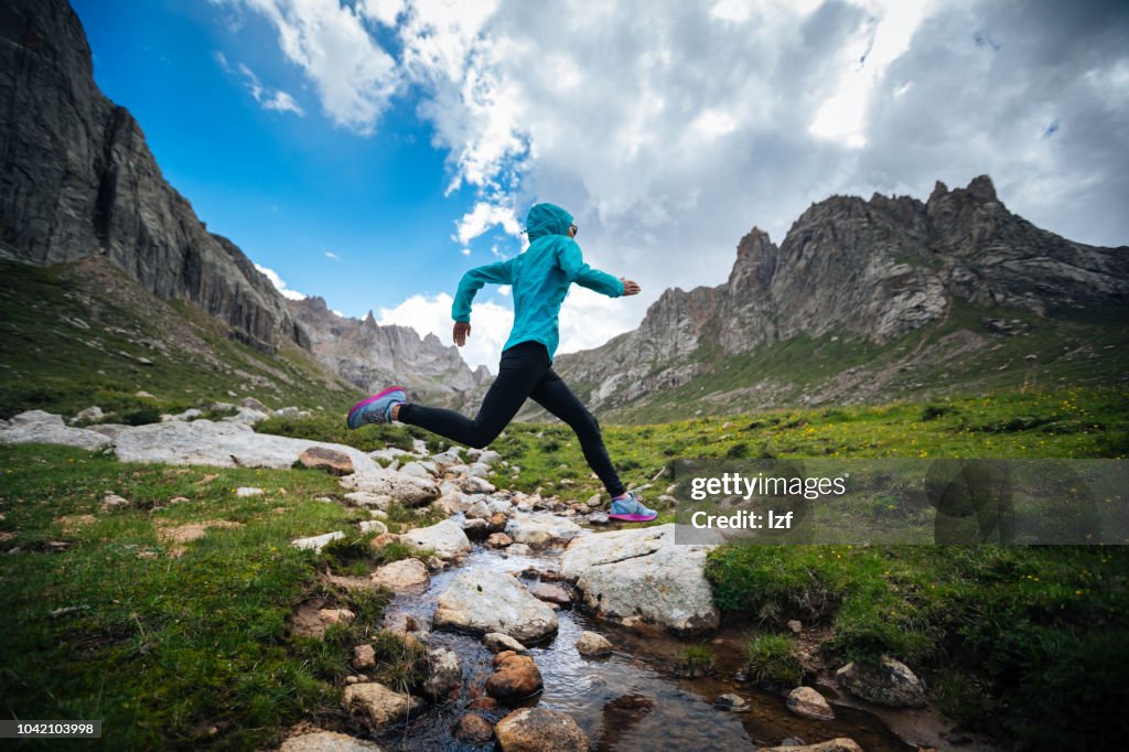 Corredor de trail de mujer saltando por encima de río del samll en montañas
