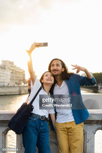 france, paris, happy young couple taking a selfie at river seine at sunset - paris millenials stock-fotos und bilder