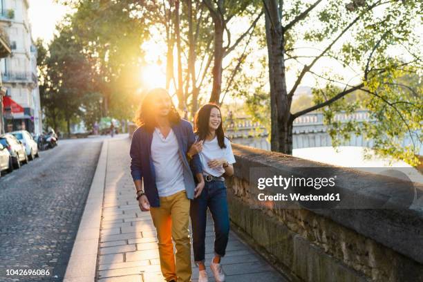 france, paris, happy young couple walking at river seine at sunset - paris street stock-fotos und bilder