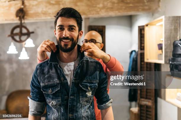 handsome man trying on new denim jacket with stylist in modern boutique - denim jacket fotografías e imágenes de stock