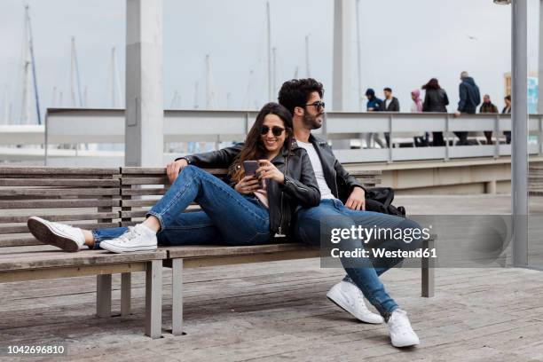 spain, barcelona, happy young couple with cell phone resting on a bench - matching outfits stock pictures, royalty-free photos & images
