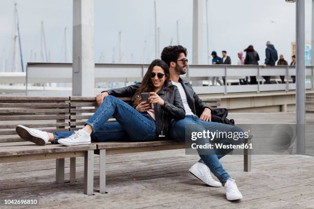 spain, barcelona, happy young couple with cell phone resting on a bench - mesma roupa imagens e fotografias de stock