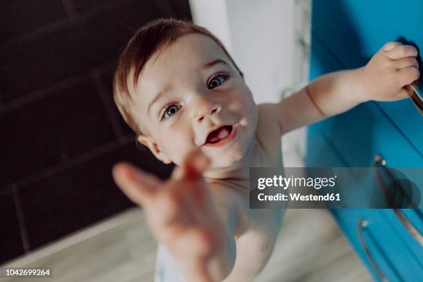 laughing baby boy holding on to chest of drawers, reaching up - baby walking stock-fotos und bilder