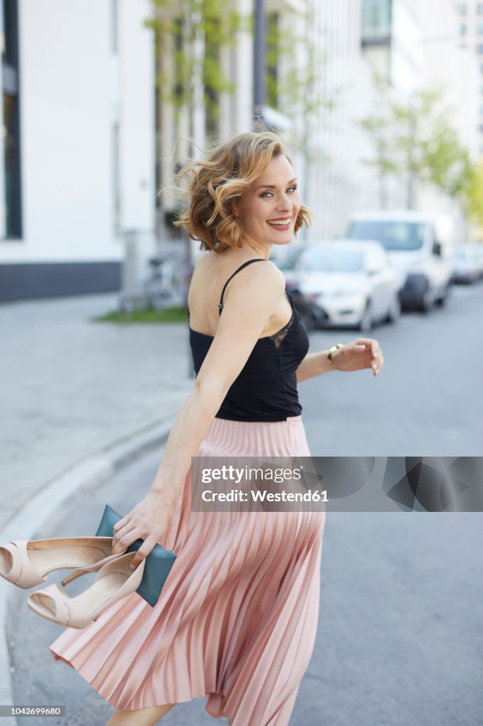 Portrait of laughing woman with high heels and clutch bag in her hand walking on the street