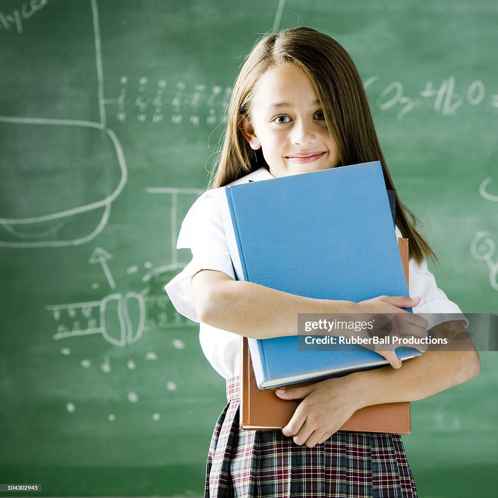 Girl in a classroom standing in front of a chalkboard holding a book