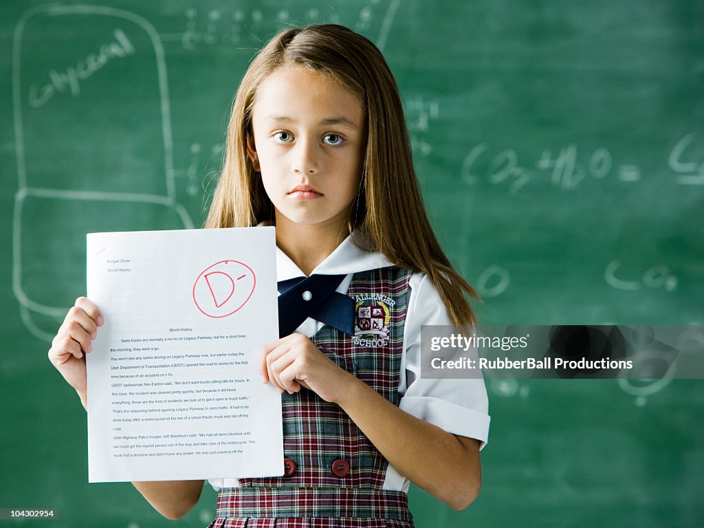 Girl in a classroom standing in front of a chalkboard holding a paper with a d minus grade
