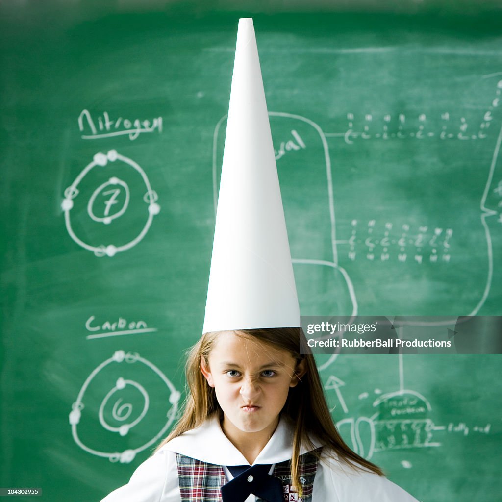 Girl in a classroom standing in front of a chalkboard wearing a dunce cap