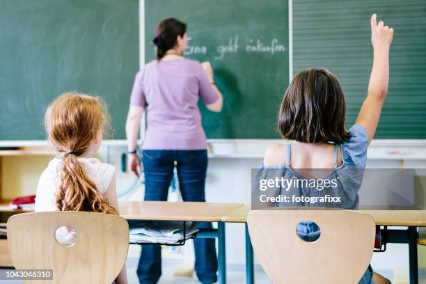 female teacher writes at the blackboard. rear view of two girls in front, on with raised arm. - northern european descent stock pictures, royalty-free photos & images