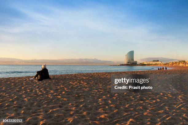 traveler woman sitting on cold beach with cup of tea with lights at sunset in barcelona, spain. - winter barcelona stock pictures, royalty-free photos & images