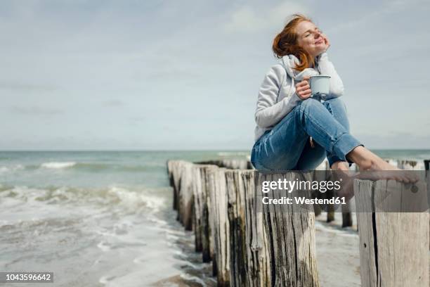 woman sitting on fence at the sea, drinking tea - barefoot redhead ストックフォトと画像
