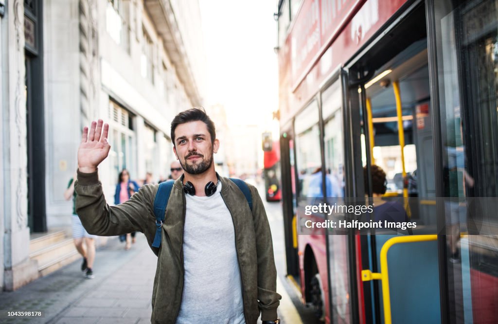 A young man with headphones standing on the bus stop, waving to somebody.