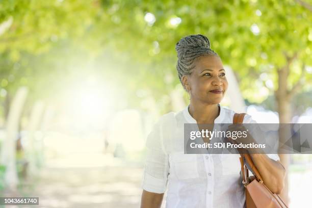 senior woman enjoys outdoor stroll on tree lined path - shoulder bag stock pictures, royalty-free photos & images