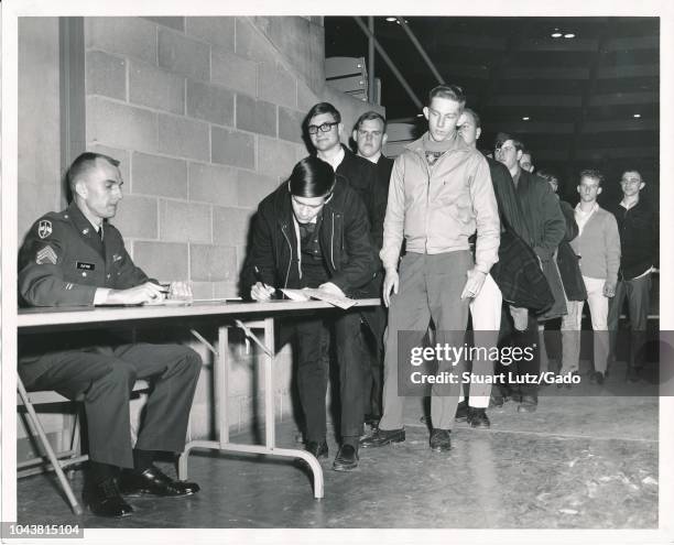 Black and white photograph of a uniformed, United States Military Officer, seated at a desk in an auditorium, watching a young man leaning over the...