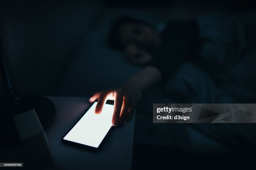 Woman reaching and switching off disturbing calls from work on smartphone while sleeping at midnight