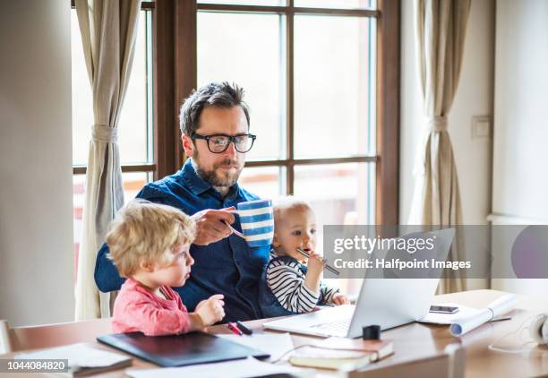 mature man with a boy and girl working at home office, using laptop and looking after his toddler children. - baby cup fotografías e imágenes de stock