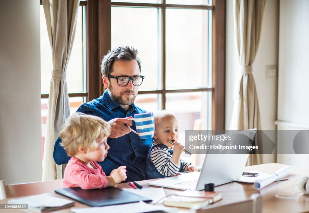 Mature man with a boy and girl working at home office, using laptop and looking after his toddler children.