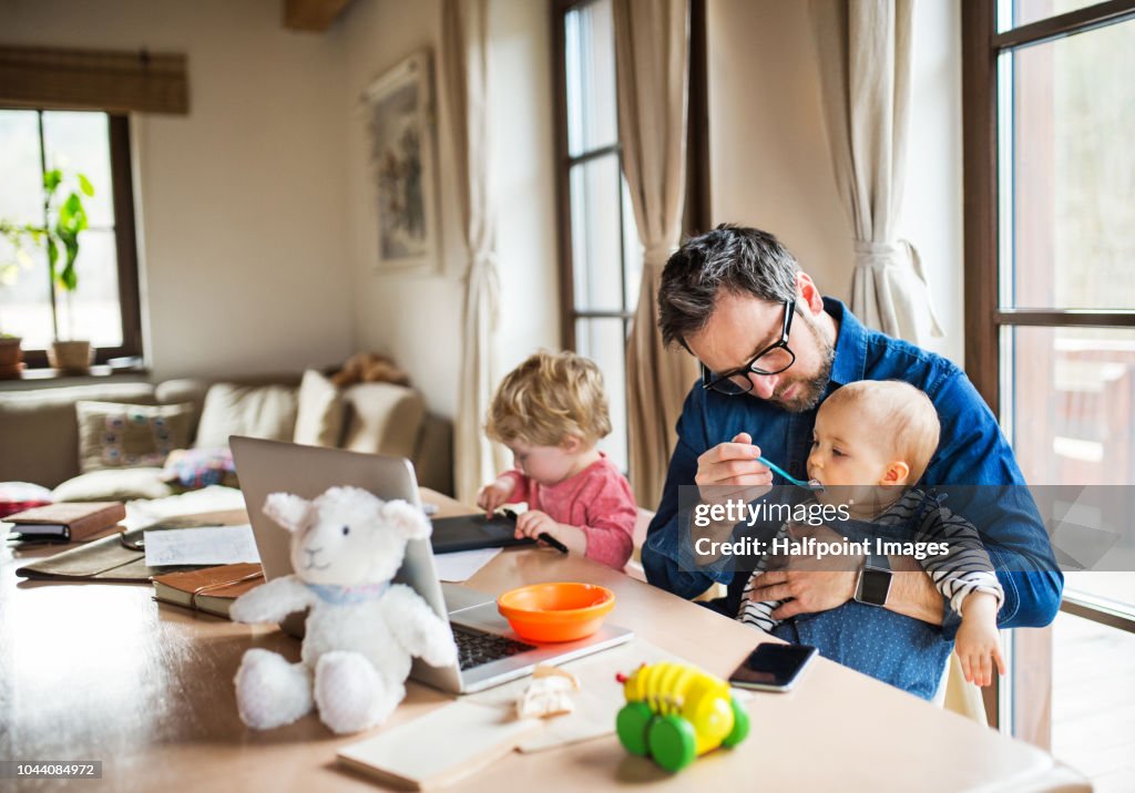 Mature man with a boy and girl working at home office, using laptop and feeding his toddler children.