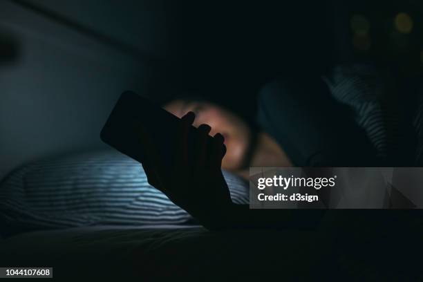 young woman text messaging on smartphone while relaxing and lying on bed at night - insomnia foto e immagini stock