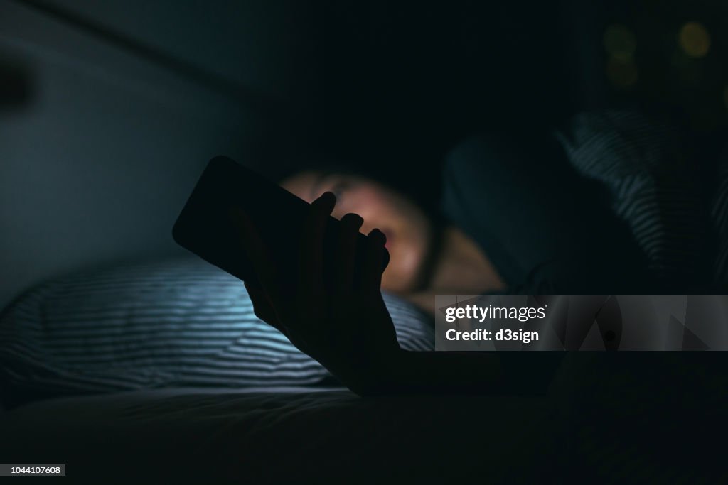 Young woman text messaging on smartphone while relaxing and lying on bed at night
