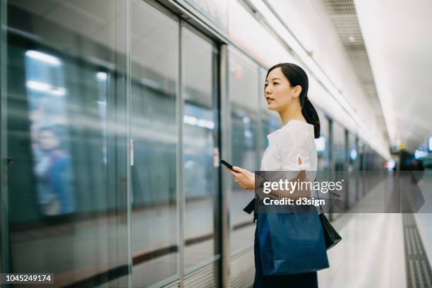 young woman carrying shopping bag using smartphone while waiting for subway in platform - un giorno nella vita foto e immagini stock