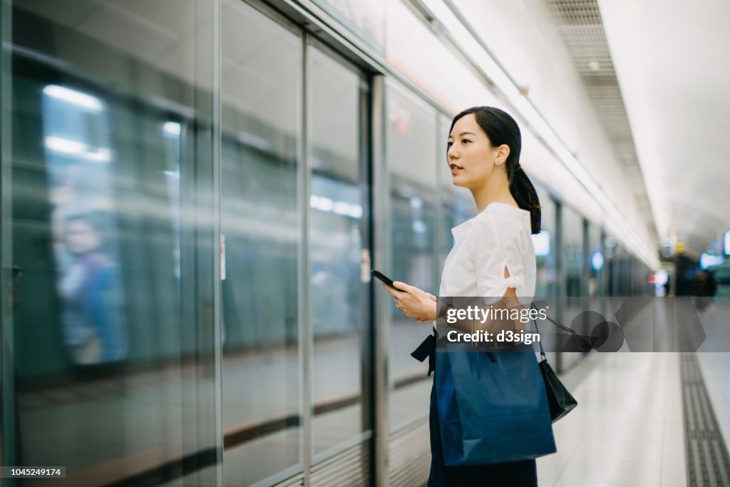 Young woman carrying shopping bag using smartphone while waiting for subway in platform