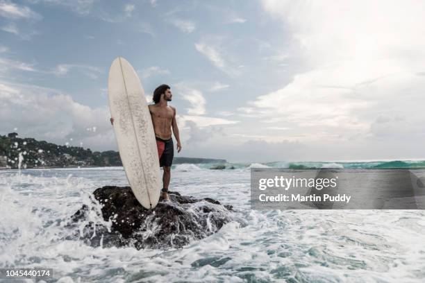 young man standing on coastline, holding a surfboard - uluwatu stock pictures, royalty-free photos & images