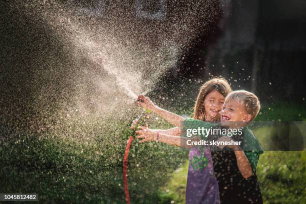 zwei verspielte kinder spaß beim spritzwasser mit gartenschlauch. - two kids playing with hose stock-fotos und bilder