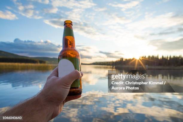 hand holding beer bottle against lake, kamloops, british columbia, canada - beer bottle stock-fotos und bilder
