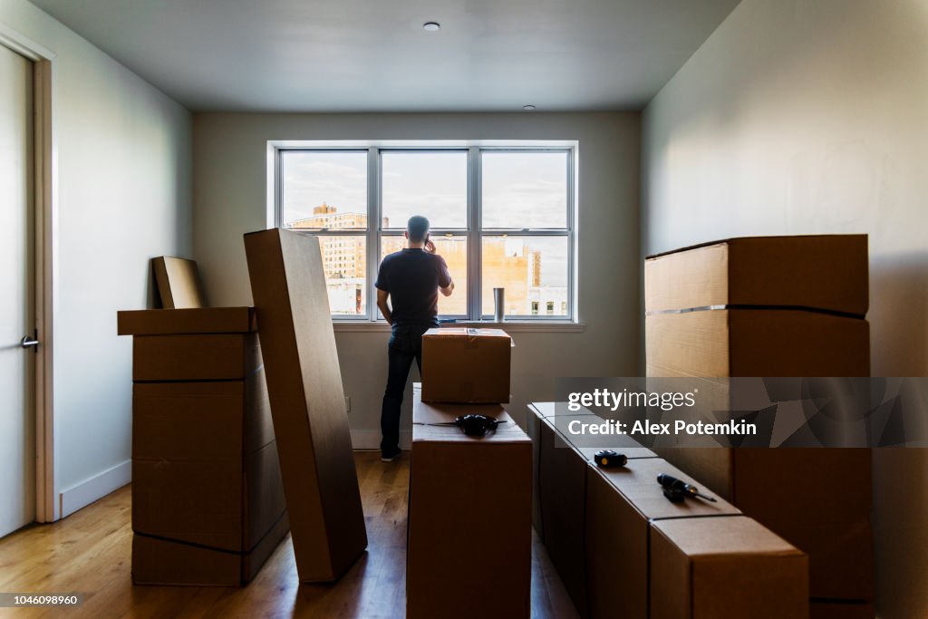 Moving in! The mature Caucasian man talking by phone nearby the window in the empty living room filled with cardboard boxes, in the new house.